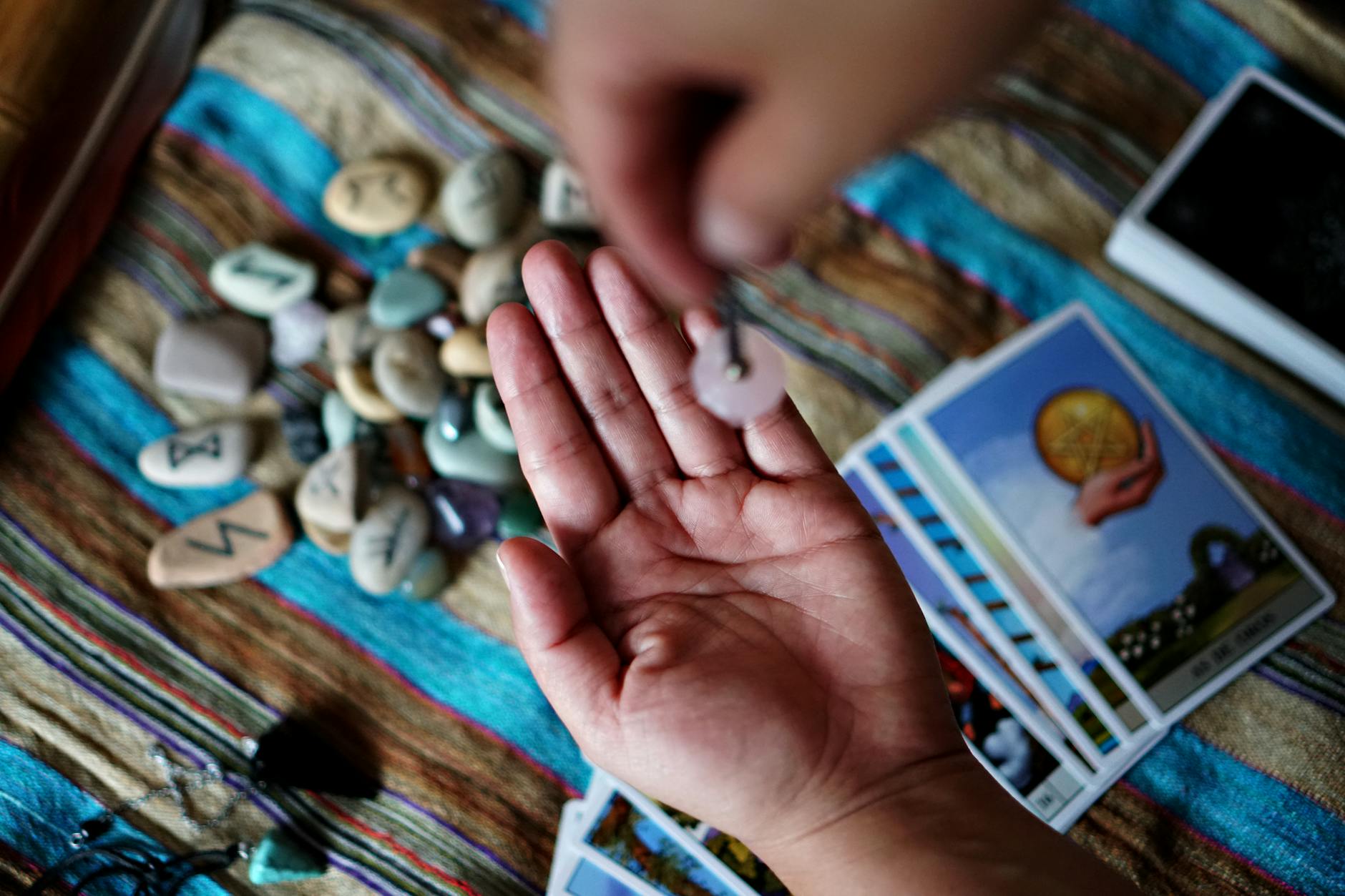 Dłoń z wahadełkiem nad runami i kartami tarota podczas wróżenia