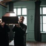 Dwaj mężczyźni w czerni niosą drewnianą trumnę w ciemnym pokoju